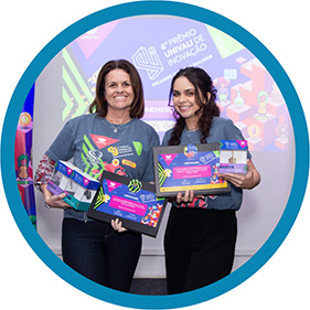 #PraTodosVerem A imagem mostra duas mulheres em pé, com prêmios nas mãos, sorrindo e posando para a foto em frente a um banner.