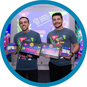 #PraTodosVerem A imagem mostra dois homens em pé, com prêmios nas mãos, sorrindo e posando para a foto em frente a um banner.