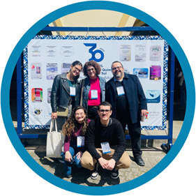 #PraTodosVerem A imagem mostra três mulheres e dois homens, duas mulheres e um homem em pé e uma mulher e um homem abaixado, sorrindo e posando para a foto em frente a um banner.